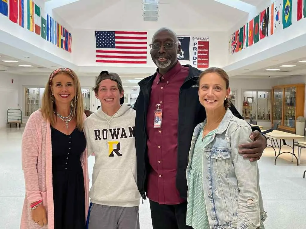 Four diverse individuals standing together in a school hallway decorated with international flags and an American flag, smiling for a photo.