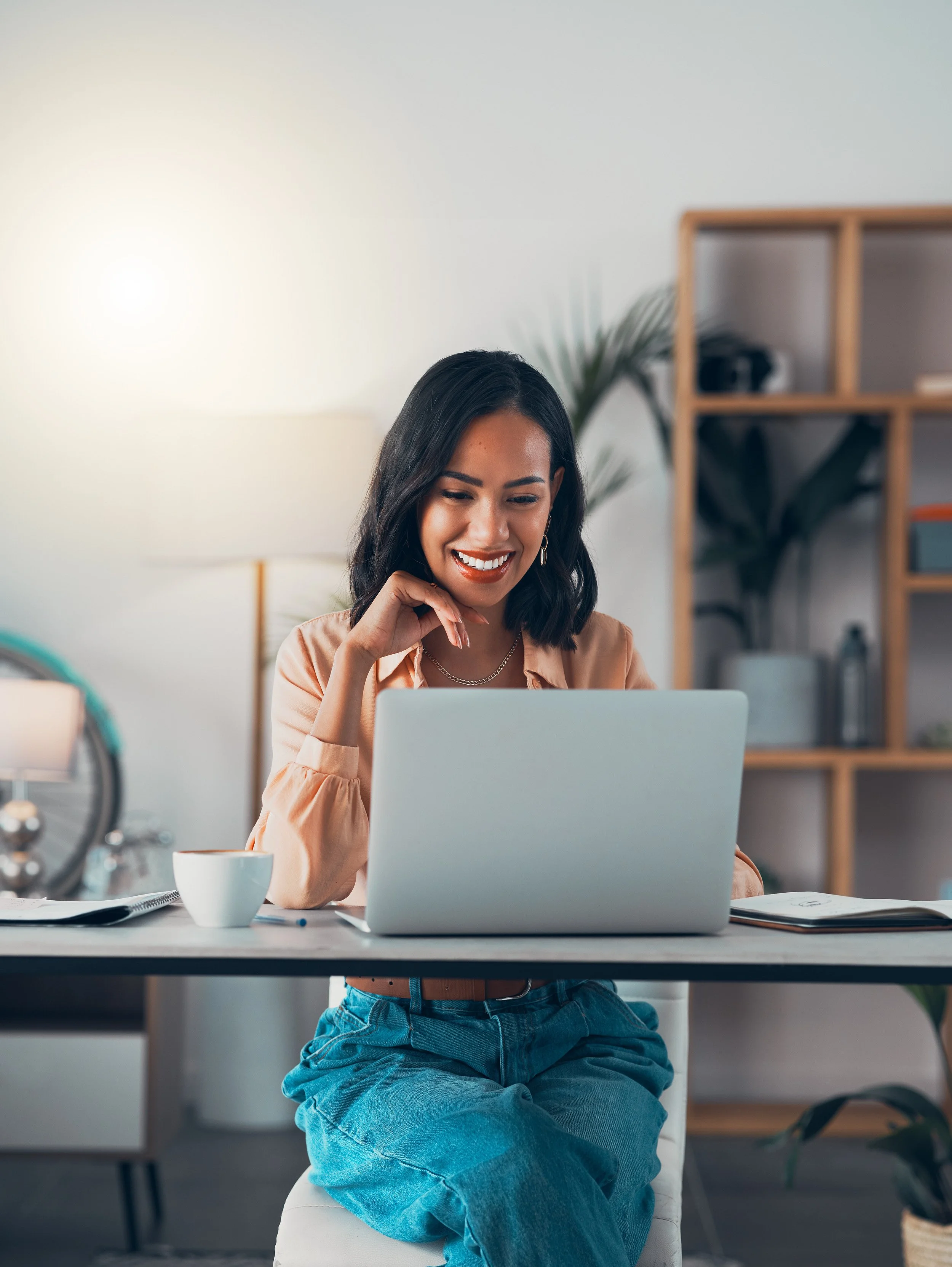A woman sitting at a desk, smiling while looking at her laptop in a bright, modern office setting.