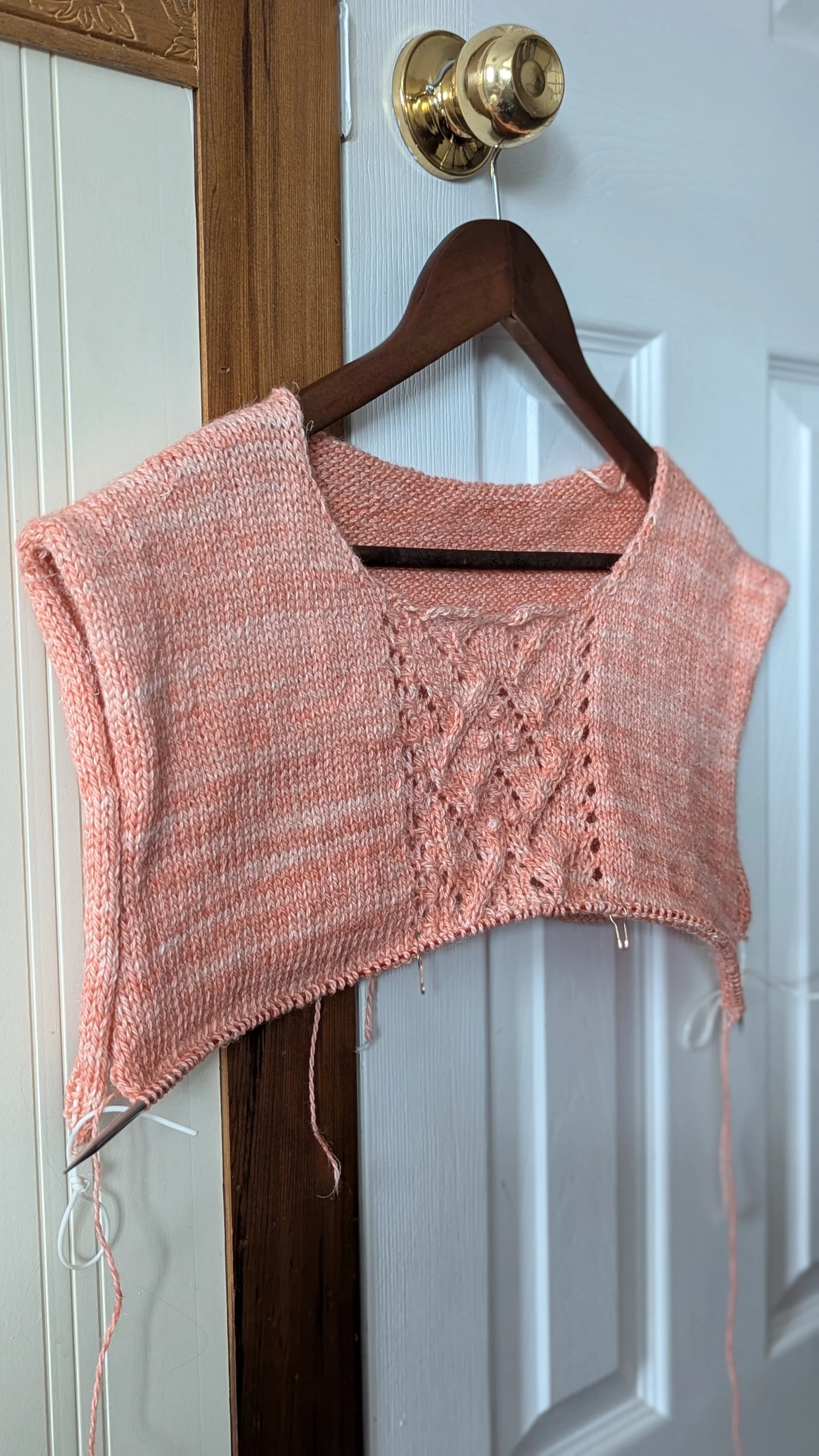 Un gilet en cours de tricotage suspendu sur un cintre en bois, accroché à une porte blanche avec une épingle à nourrice en bas.