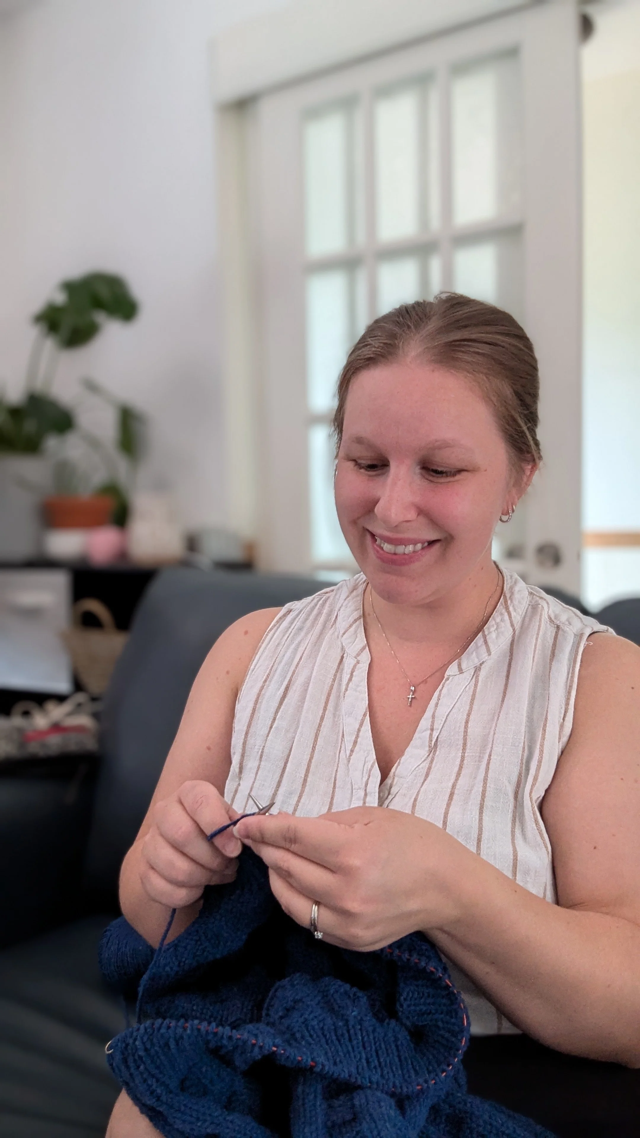 Femme souriante qui tricote avec du fil bleu dans un salon lumineux.