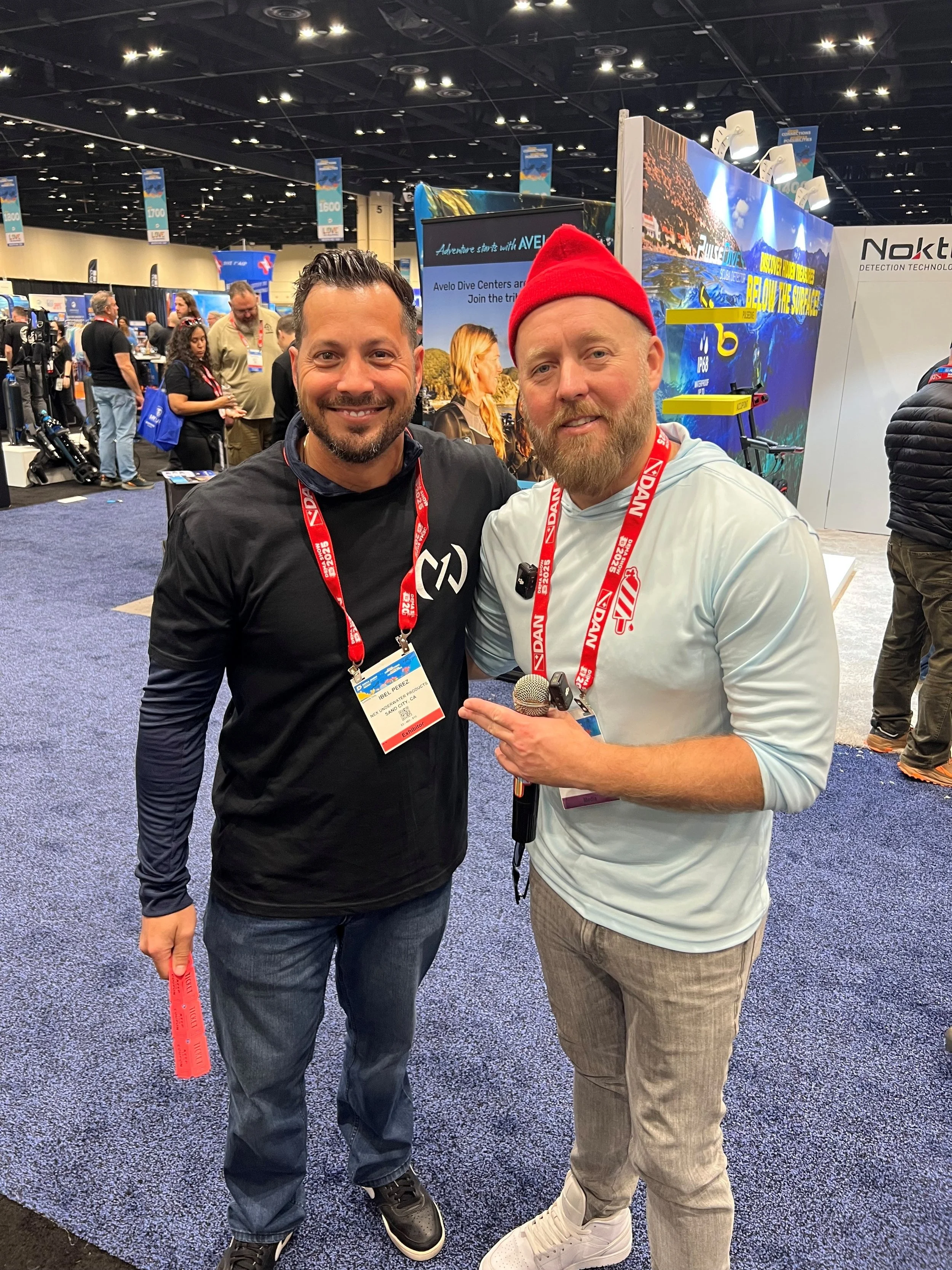 Two men at a conference or trade show, one wearing a black long-sleeve shirt and the other in a white hoodie with a red beanie. They are smiling and standing close together, with the man in white pointing to the other man. The background shows other attendees, booths, and displays.