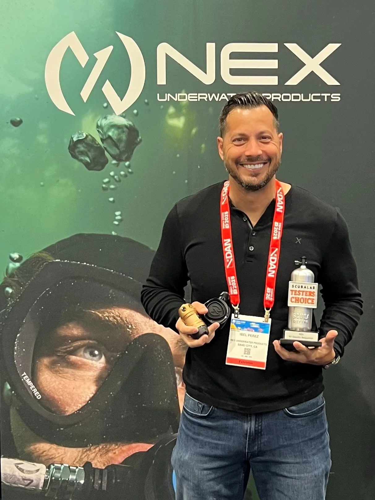 A man smiling and holding awards, standing in front of a NEX underwater products display at a conference, wearing a black shirt and a red conference badge.