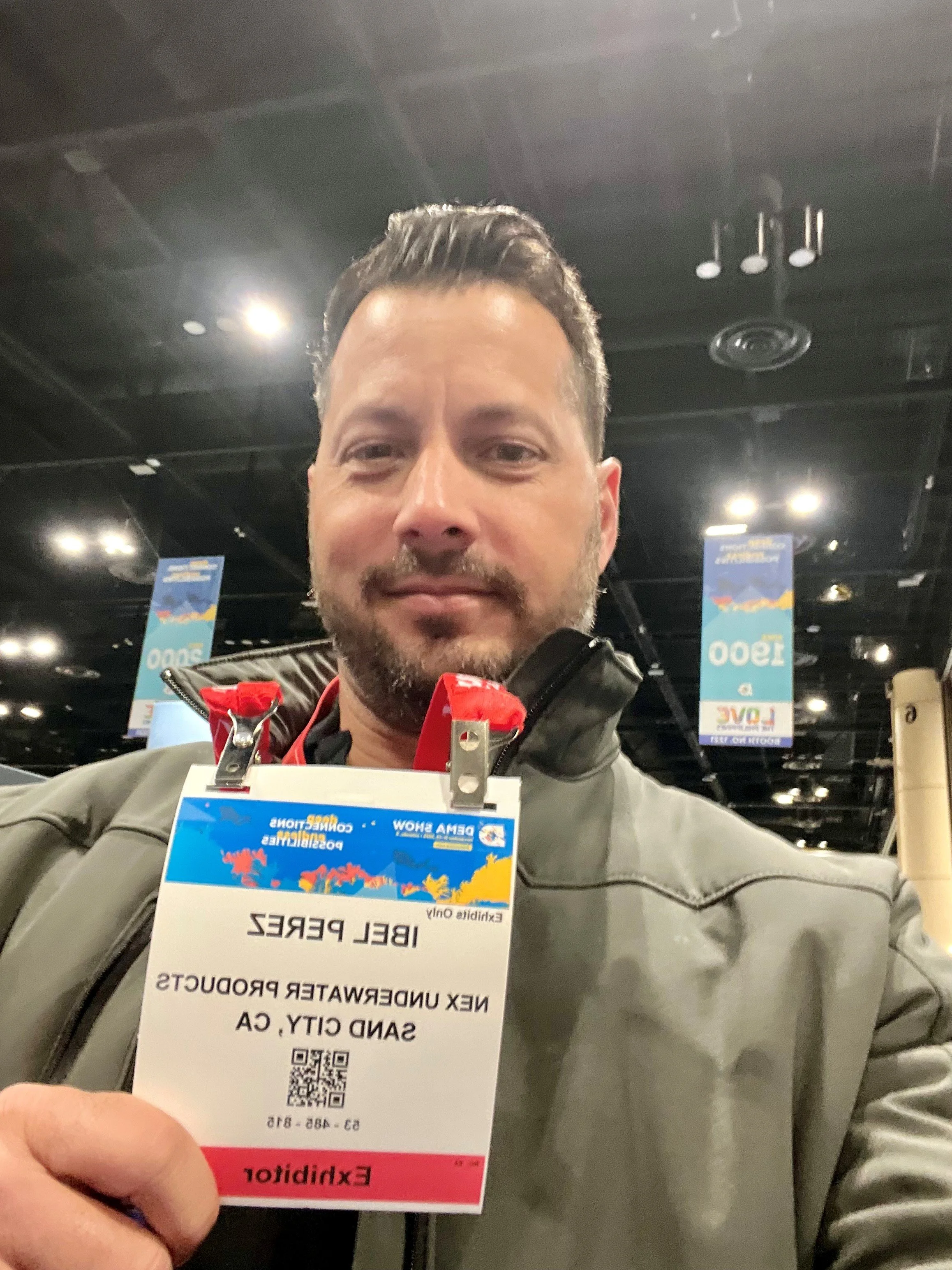 A man at an expo shows a 2025 DEMA badge that reads 'IBEL PEREZ NY NEX UNDERWATER PRODUCTS SAND CITY, CA.' He is smiling slightly and wearing a green jacket, with a large convention hall in the background.