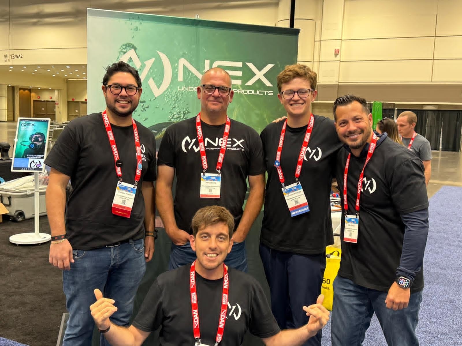 Group of five men wearing black T-shirts with 'NEX' logo and name tags, smiling at a conference, standing in front of a green 'NEX' banner, with one man kneeling in front giving a thumbs up.