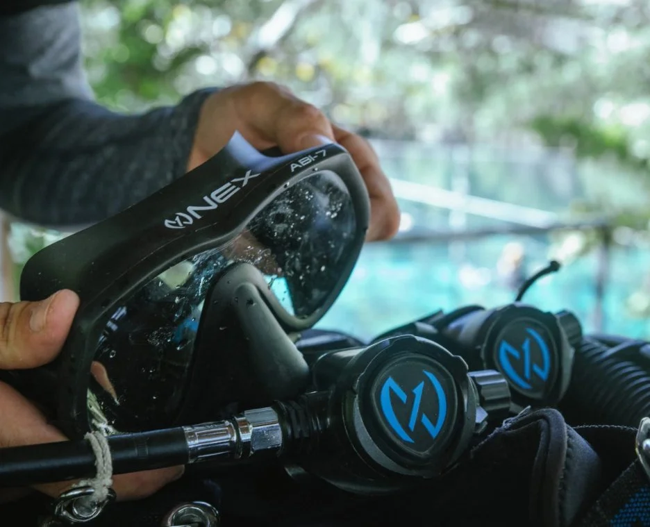 A person holding a black diving mask and two scuba regulators on a surface, with a blurred outdoor background.