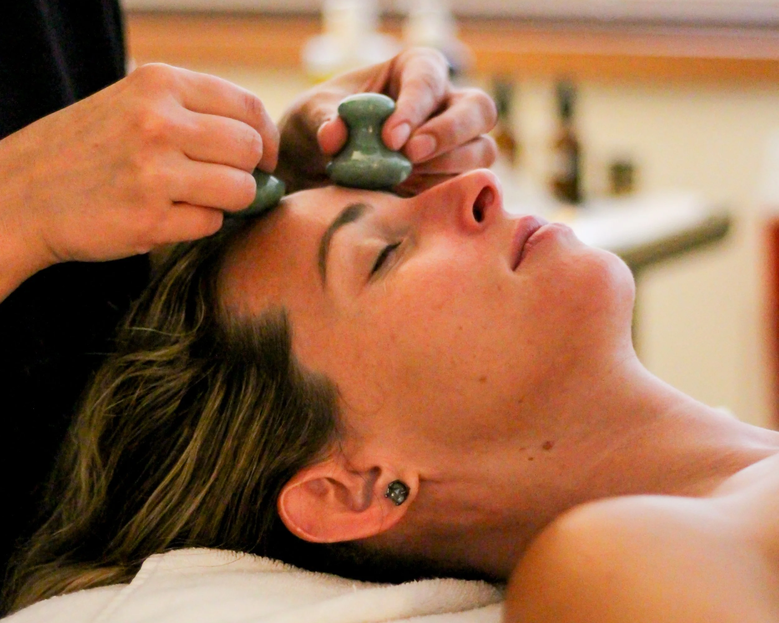 A woman lying on a treatment bed with her eyes closed, receiving a facial treatment with a facial mask being applied by a therapist using a brush.