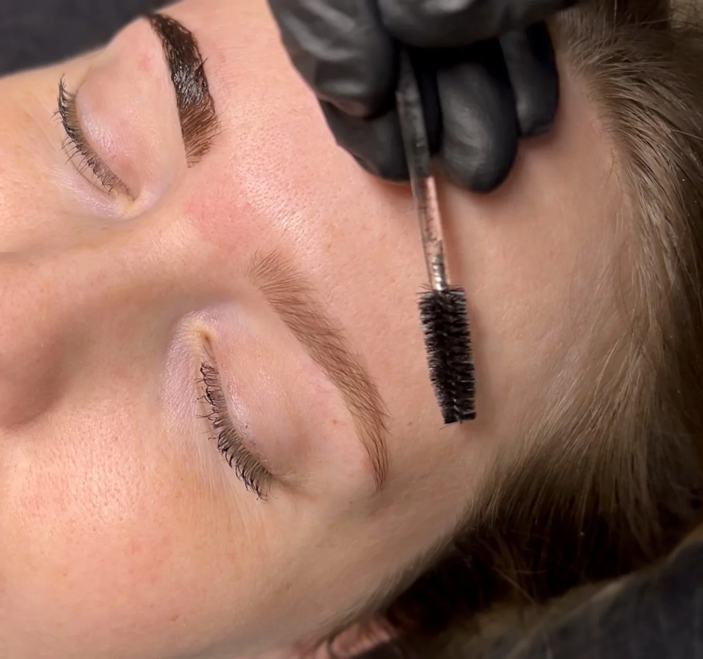 A person lying down with eyes closed, receiving a cosmetic eyebrow treatment using a tool held by a gloved hand.