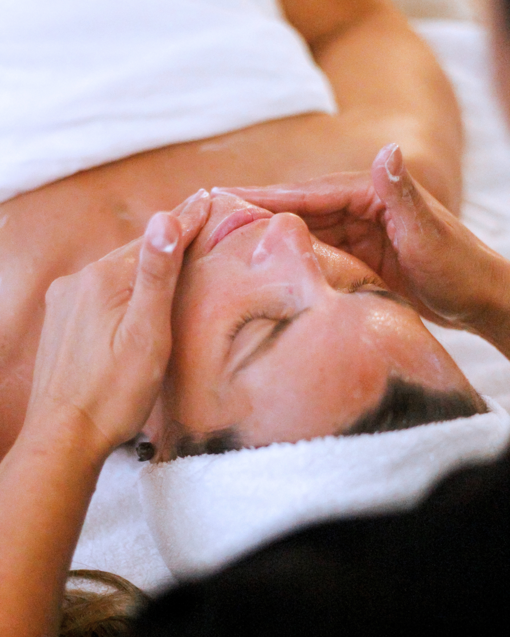 Person receiving facial massage with closed eyes, lotion or mask applied on face, massage therapist's hands gently touching face.