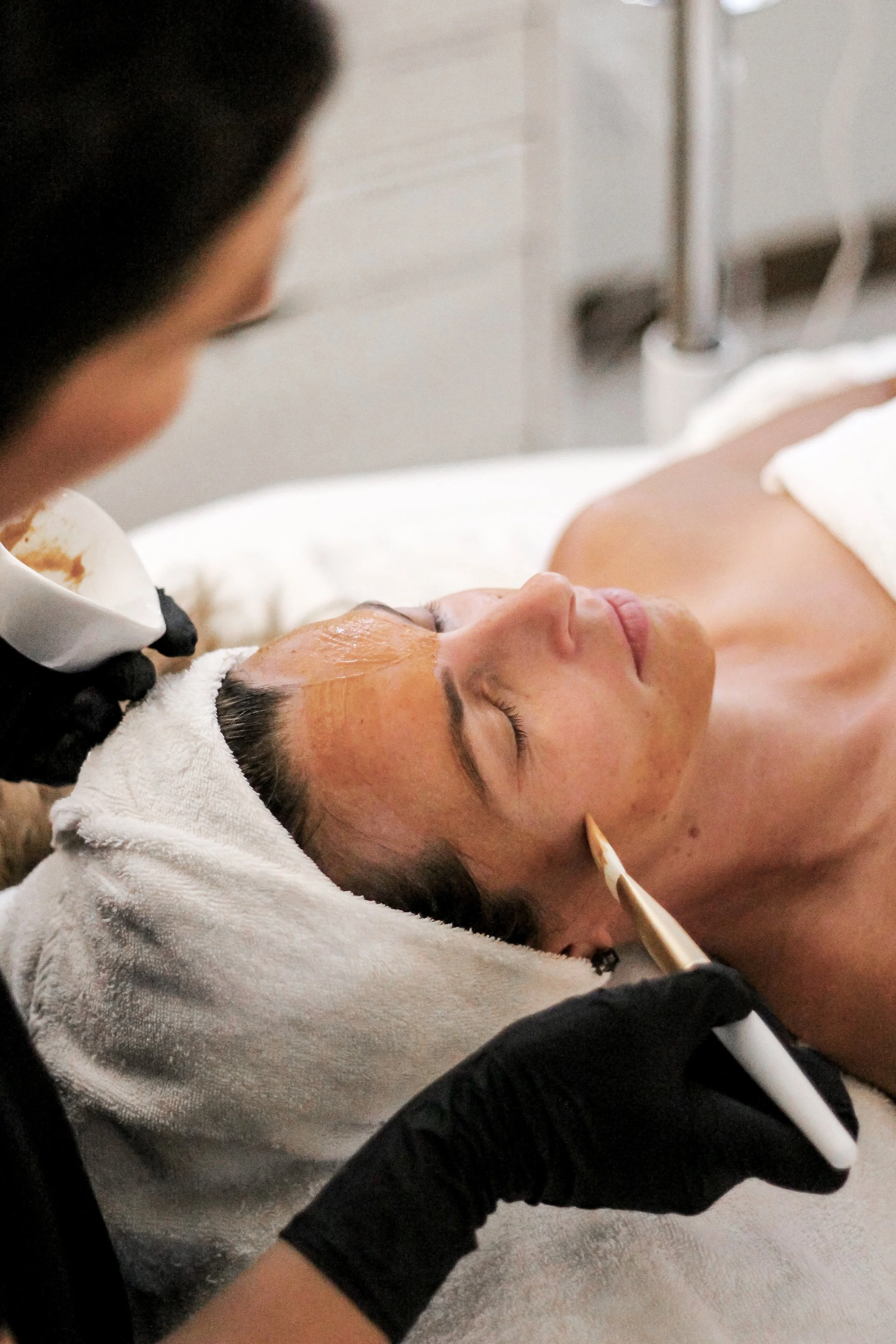 A woman receiving a facial massage while lying on her back on a white massage table, covered with a white towel around her waist.