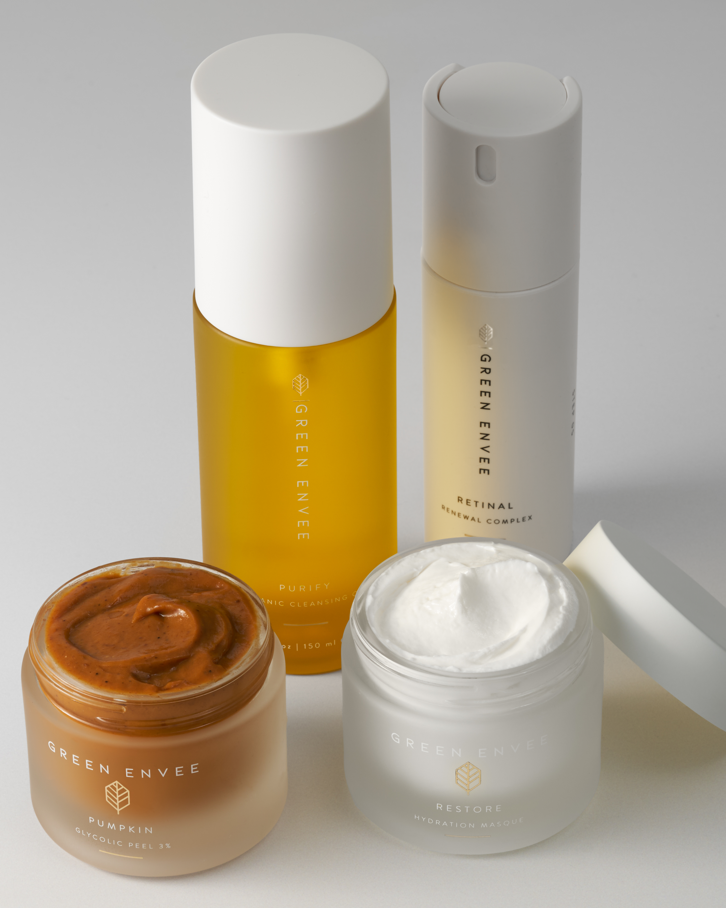 Skincare products from Green Envee, including a white and yellow bottle, a white pump bottle, and two open jars of cream, one with a brownish-orange peel mask and the other with a white hydrating masque.