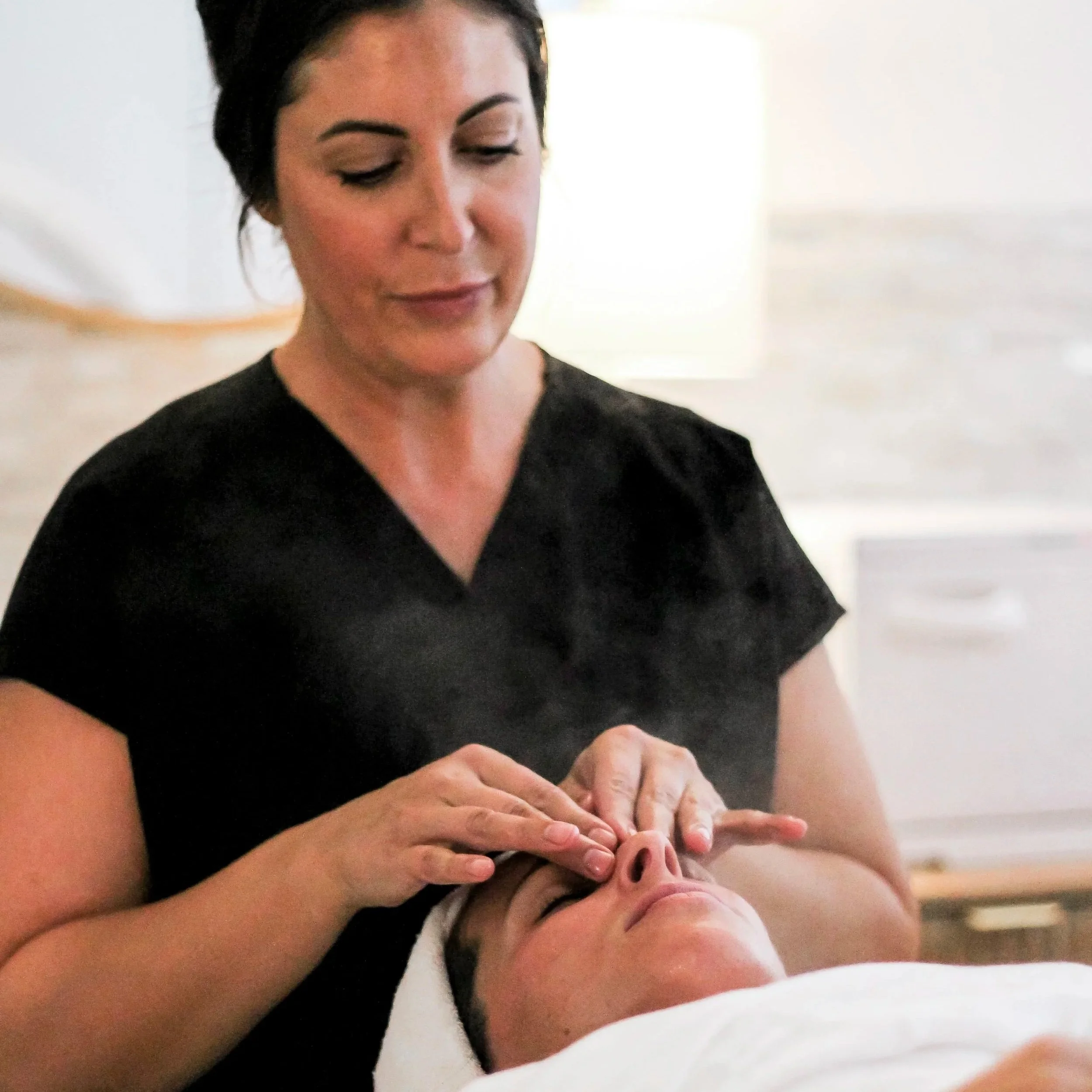 A woman lying on a treatment bed with her eyes closed, receiving a facial treatment with a facial mask being applied by a therapist using a brush.