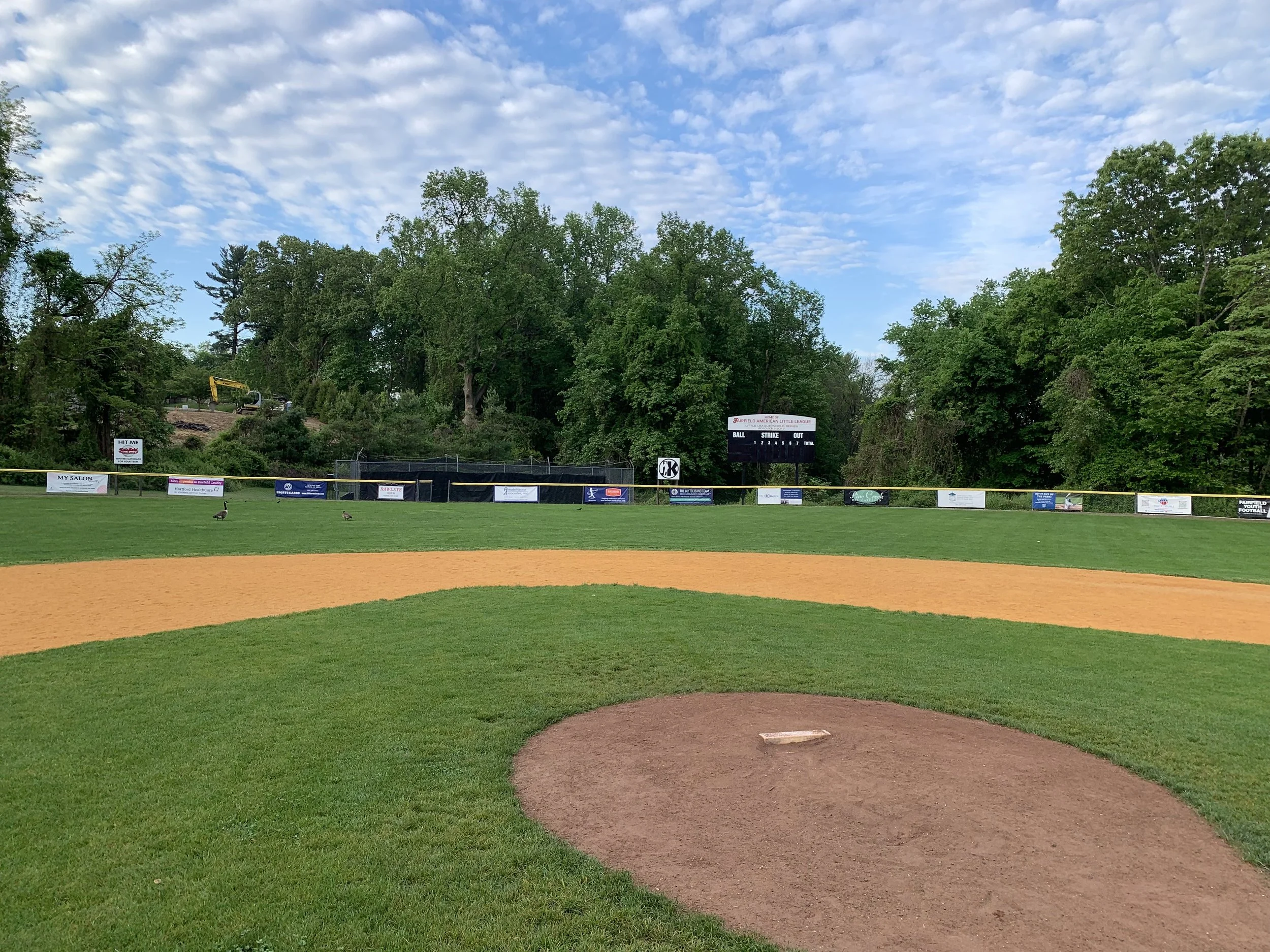 Empty baseball field with a view of the outfield, scoreboard, and surrounding trees under a partly cloudy sky.