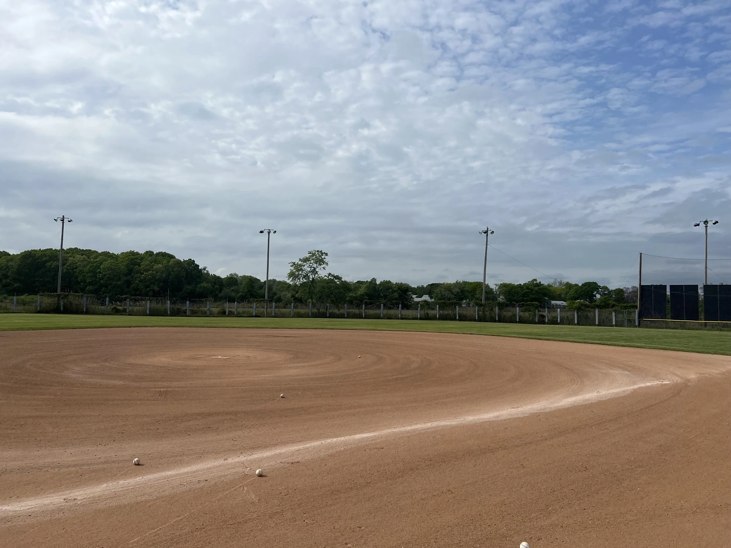 Empty baseball field with dirt infield, grass outfield, and cloudy sky overhead.