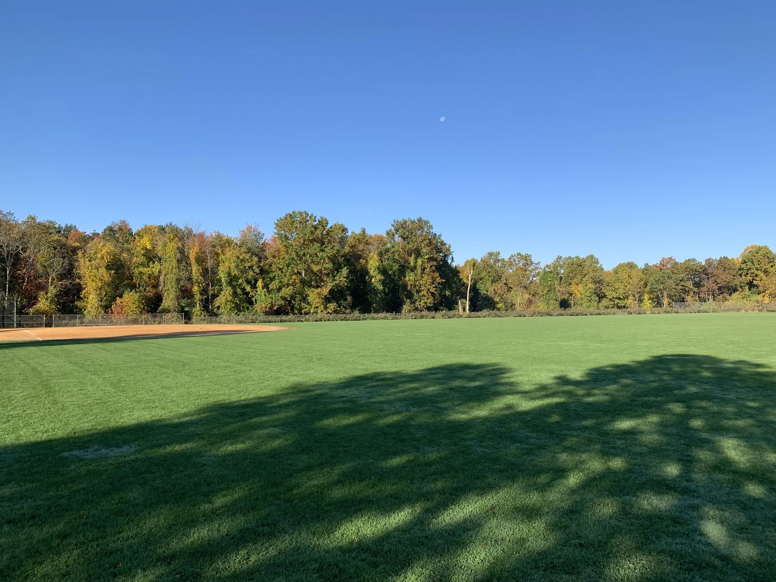An empty baseball field with green grass, trees with fall foliage in the background, and a clear blue sky with the moon visible.
