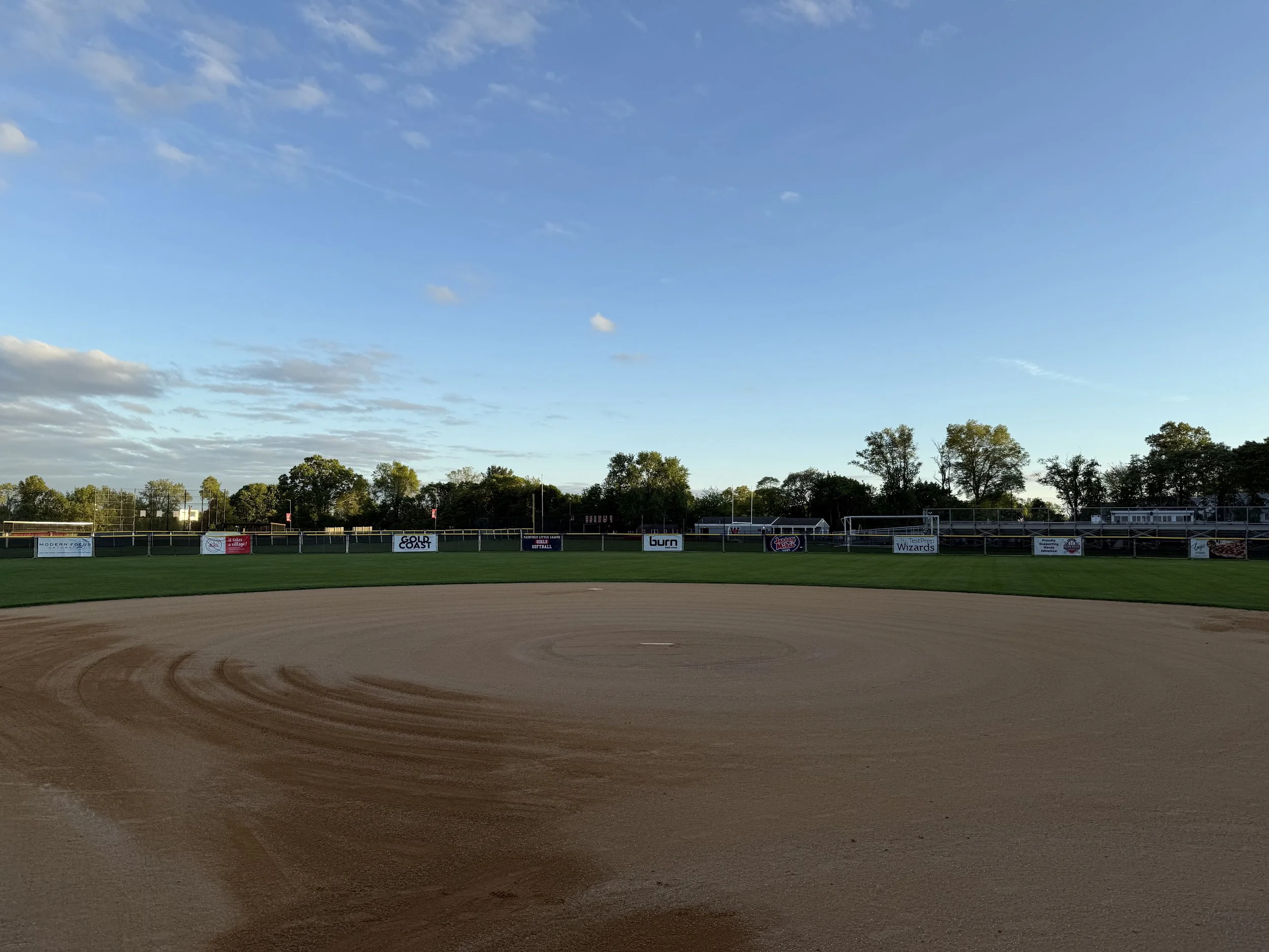 Empty baseball field with dirt infield and green outfield, surrounded by advertising banners and a tree line, under a clear sky.