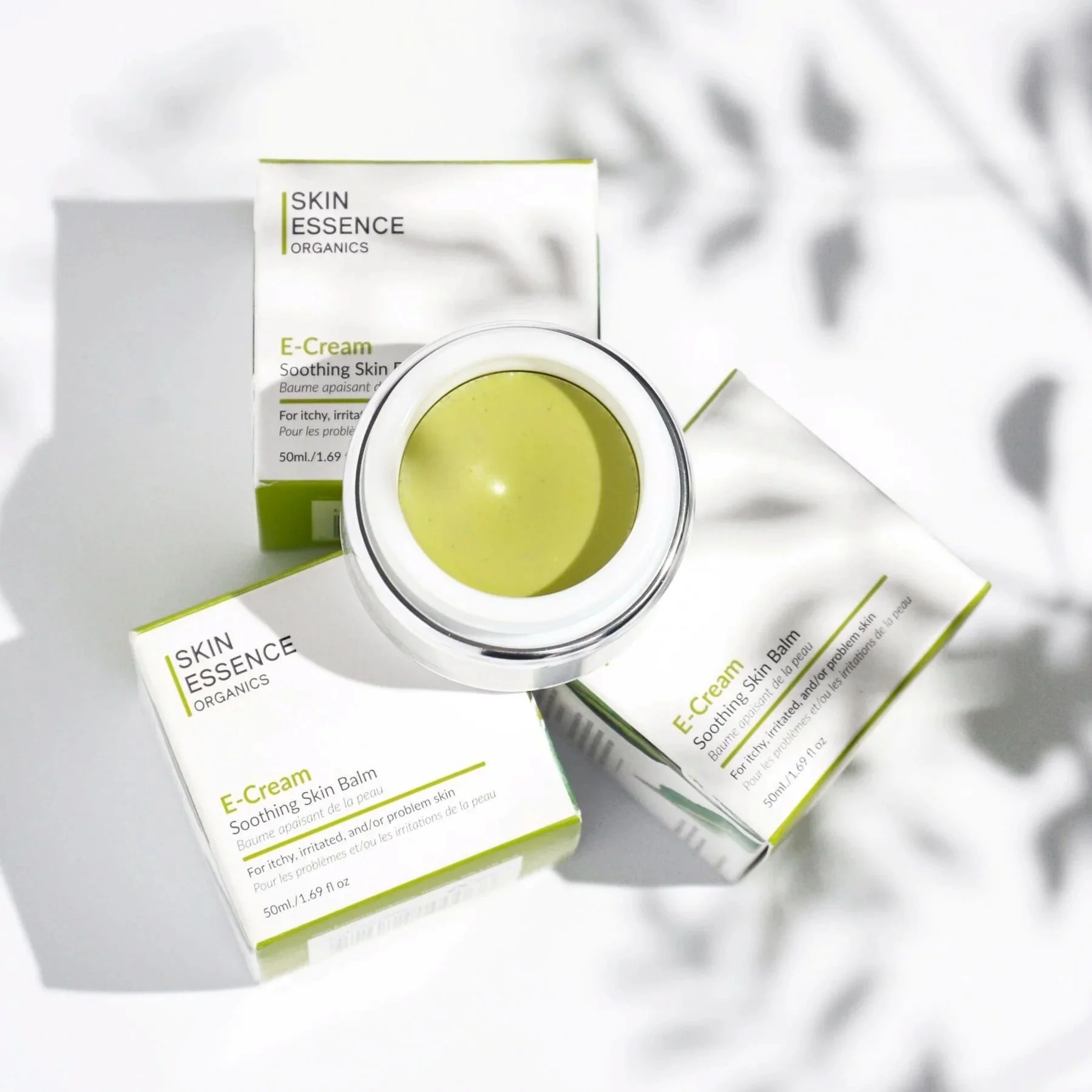 Open jar of green-colored skincare cream surrounded by three boxes of Skin Essence Organics E-Cream on a white surface with shadows.
