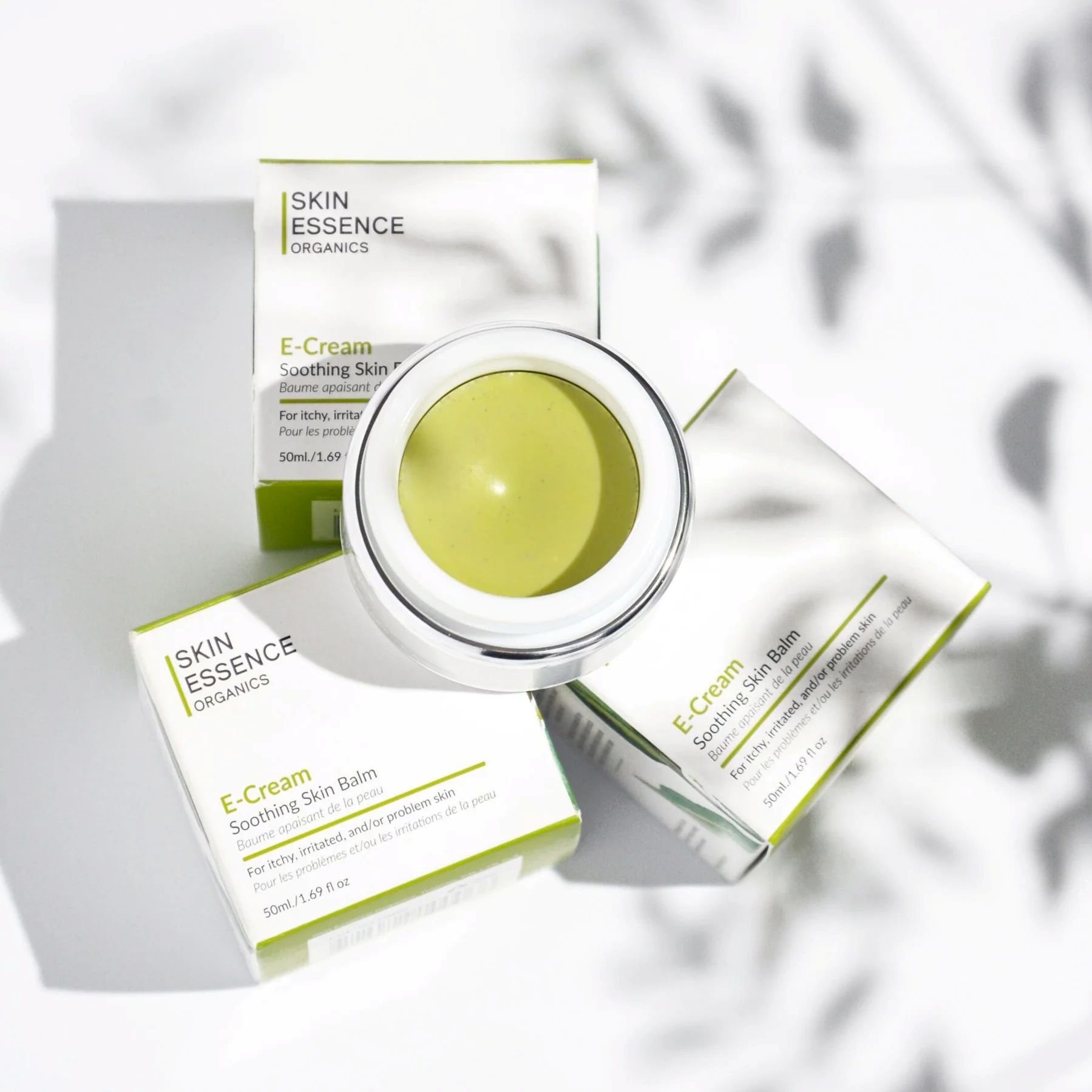 Open jar of green E-Cream soothing skin balm surrounded by three boxes of the same product, with shadows on a white surface.