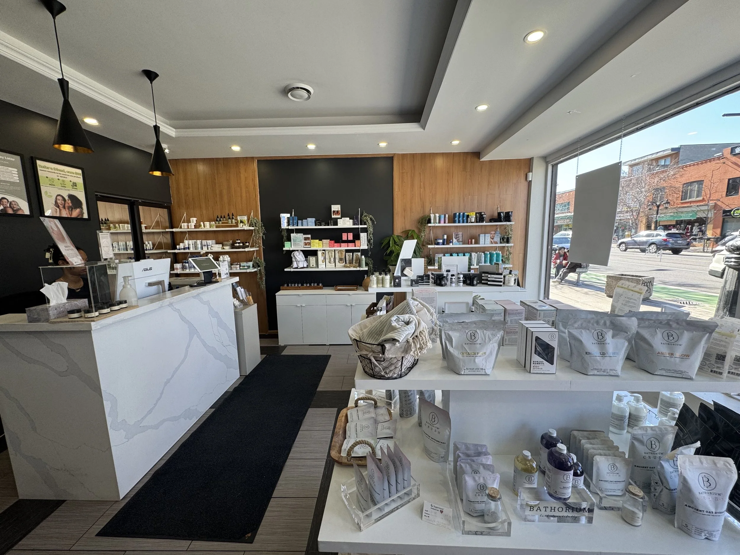 Interior of a retail store selling skincare and beauty products, with shelves stocked with various bottles, boxes, and jars, a prominent white checkout counter, and large front windows showing cars and buildings outside.
