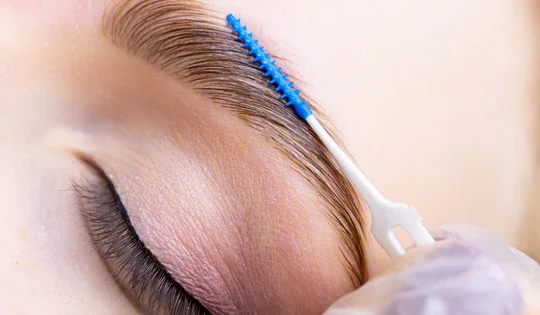 Close-up of a person with eye makeup, using a blue spoolie brush to groom their eyebrow.
