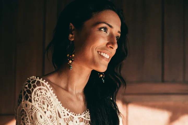 A woman with long dark hair smiling in a warmly lit room, wearing a cream-colored, patterned top and dangling earrings.
