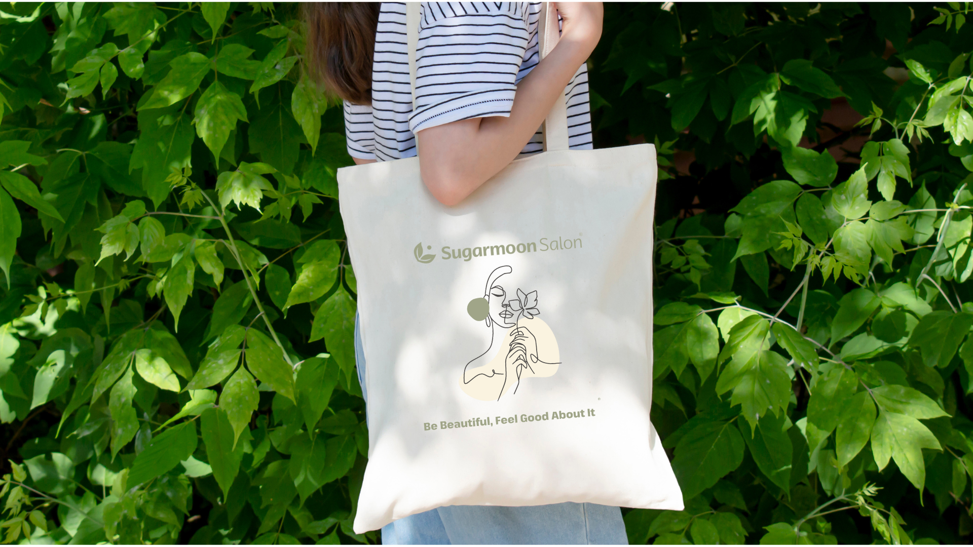 Person holding a tote bag with 'Sugarmoon Salon' logo and a line drawing of a woman with a flower, against a backdrop of green leafy plants.