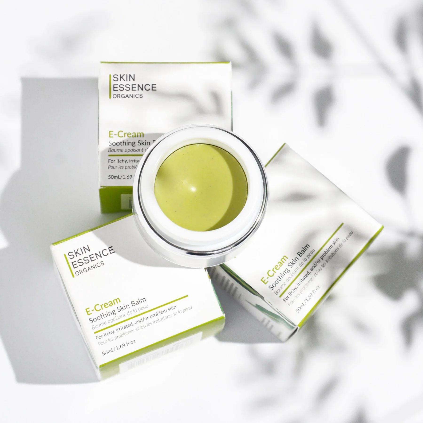 Open container of green soothing skin balm surrounded by three white and green boxes labeled Skin Essence Organics E-Cream for itchy, irritated skin.