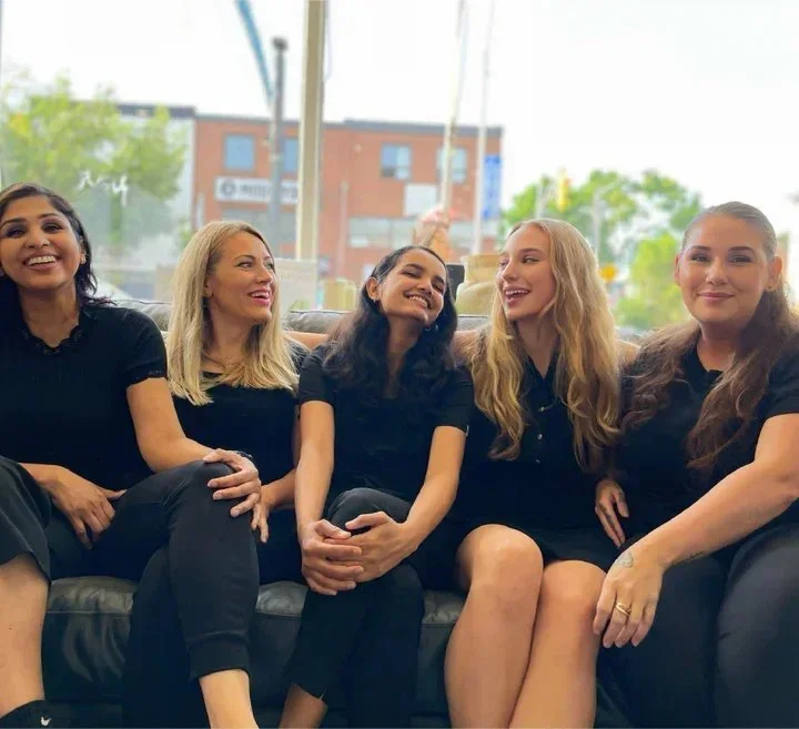 Five women sitting on a couch, smiling and laughing indoors with a window showing a cityscape in the background.
