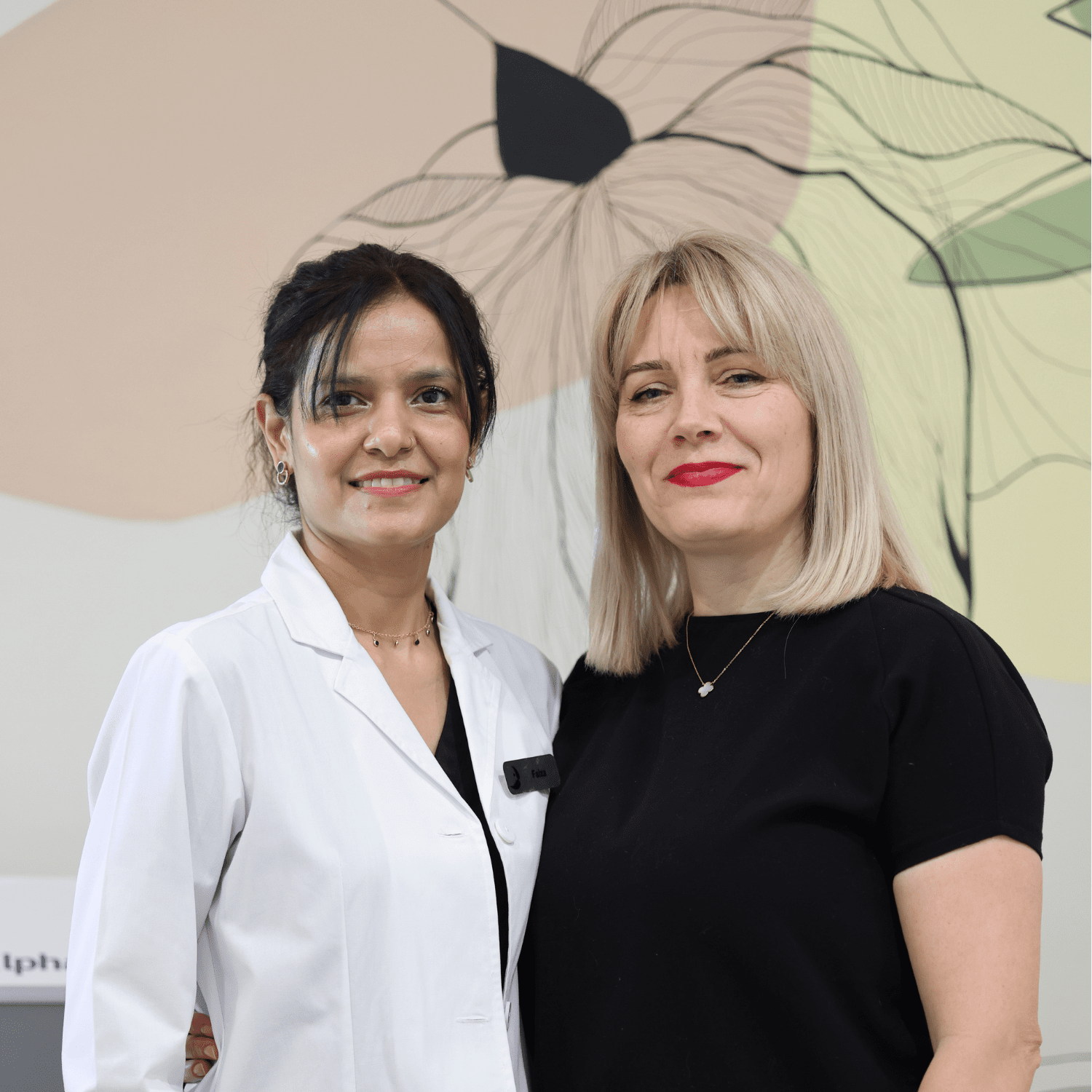 Two women standing side by side indoors, smiling, with a large abstract floral artwork in the background. One woman is wearing a white lab coat, and the other is dressed in a black top.