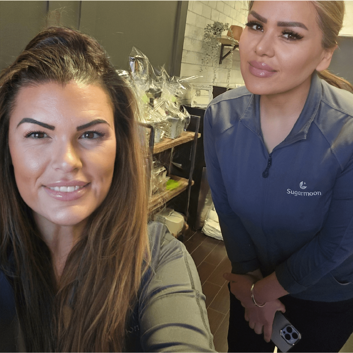 Two women are smiling and posing for a photo in a store or cafe. One woman has long brown hair and the other has light brown hair tied back, wearing a dark blue jacket with the logo 'Sugarmoon' on it. There are make-shift gift baskets wrapped in plastic behind them.