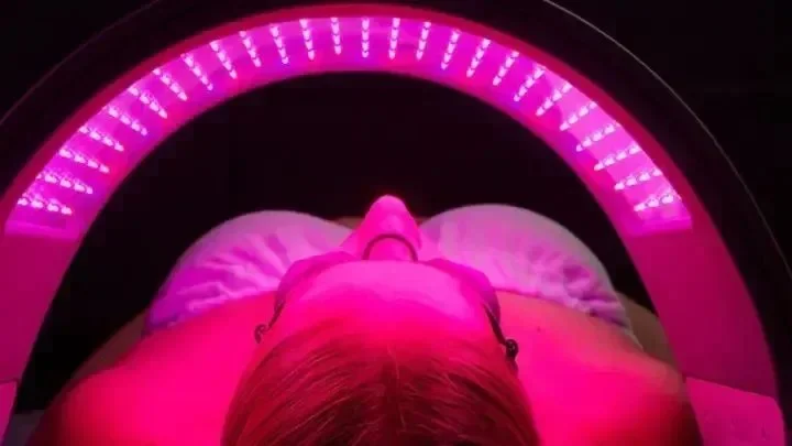 Person lying down in a pink LED light therapy bed with their eyes closed, facing upwards.
