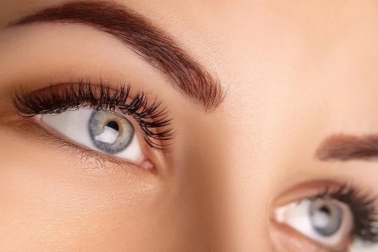 Close-up of a woman's eye, showing long eyelashes, well-defined eyebrows, and light-colored iris.