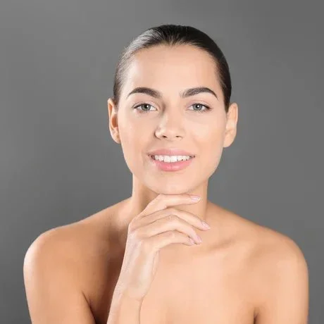 A young woman with clear skin and dark hair tied back, smiling gently at the camera against a gray background.