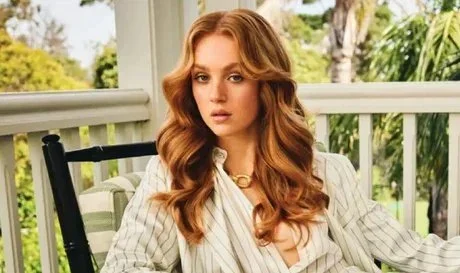 A young woman with red hair sitting on a porch with trees and plants in the background, wearing a white striped shirt and a gold necklace.