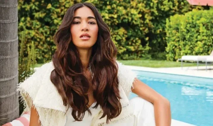 A woman with long wavy brown hair wearing a white outfit, sitting outdoors near a swimming pool with greenery in the background.