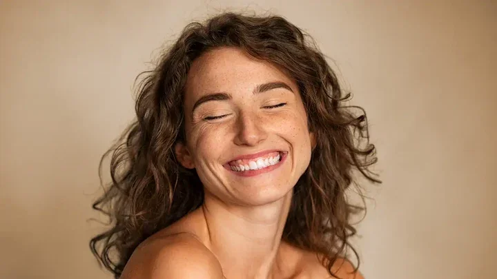 Close-up of a smiling woman with curly brown hair and freckles, with eyes closed, against a beige background.