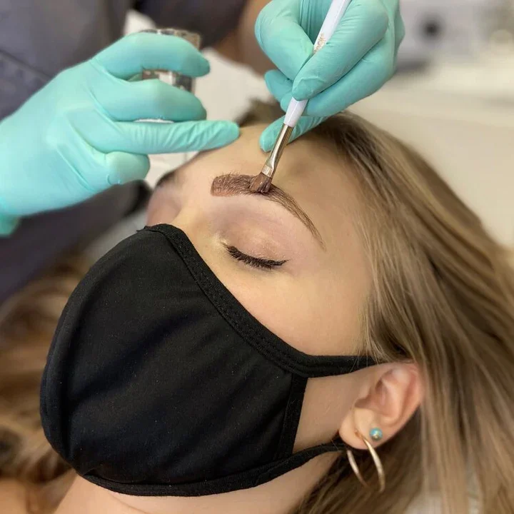 A person with a black face mask and earrings receives a microblading or eyebrow tattooing treatment from a technician wearing teal gloves.