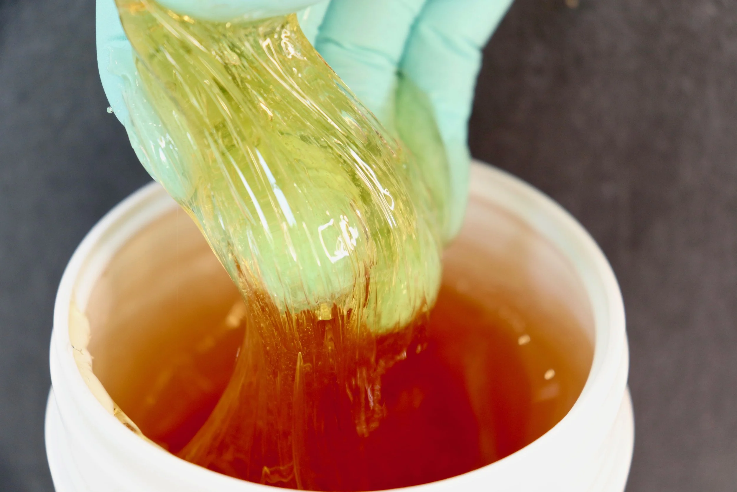 A gloved hand holding a clear plastic spatula with amber-colored honey above a white container filled with honey.