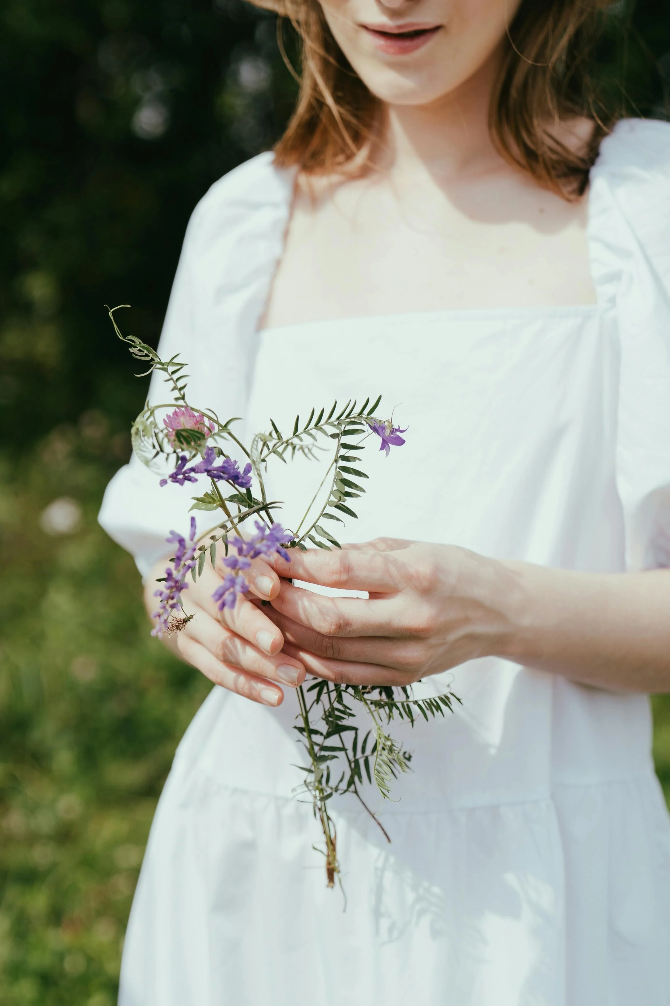 Woman in a white dress holding a small bouquet of purple and pink wildflowers outdoors.