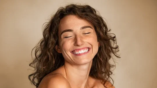 A young woman smiling with her eyes closed, showing her teeth, and with curly brown hair against a neutral background.
