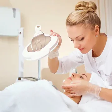 A woman in a medical uniform performs a facial treatment on a patient in a clinical setting.