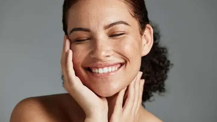 A female model with curly hair tied back, smiling with her eyes closed and hands on her chin, showcasing natural beauty
