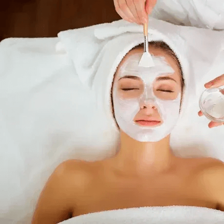 Woman receiving a facial treatment, lying with eyes closed, while a beauty professional applies a white facial mask.