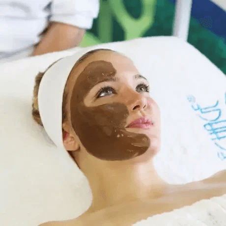 Woman lying on an aesthetic treatment bed with a mud mask applied to her face, wearing a white headband, in a spa or skincare clinic.