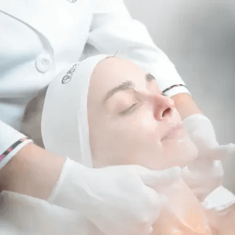 A woman receiving a facial treatment in a spa or clinic setting, lying down with her eyes closed, wearing a headband, while a professional applies skincare products or a mask.