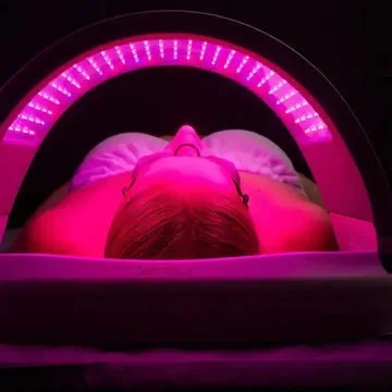 Person lying down inside a pink-lit tanning bed or light therapy device.