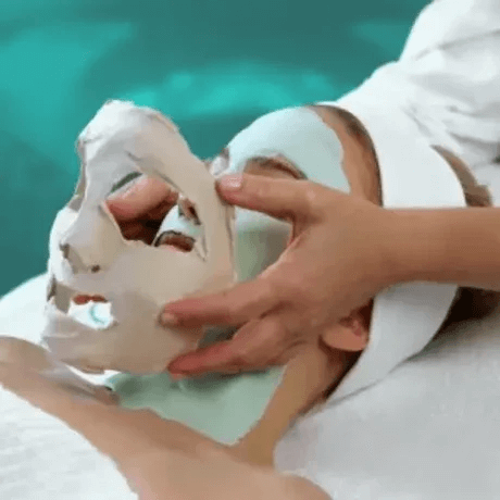 A person lying down receiving a facial treatment with a clay or mud mask, while a therapist removes the mask with their hands.