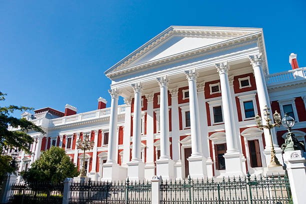 The White House is a large, historic mansion with tall white columns and a red brick facade. It features a prominent portico supported by multiple columns, decorative white trim, and is surrounded by a black metal fence. The clear blue sky is visible in the background.