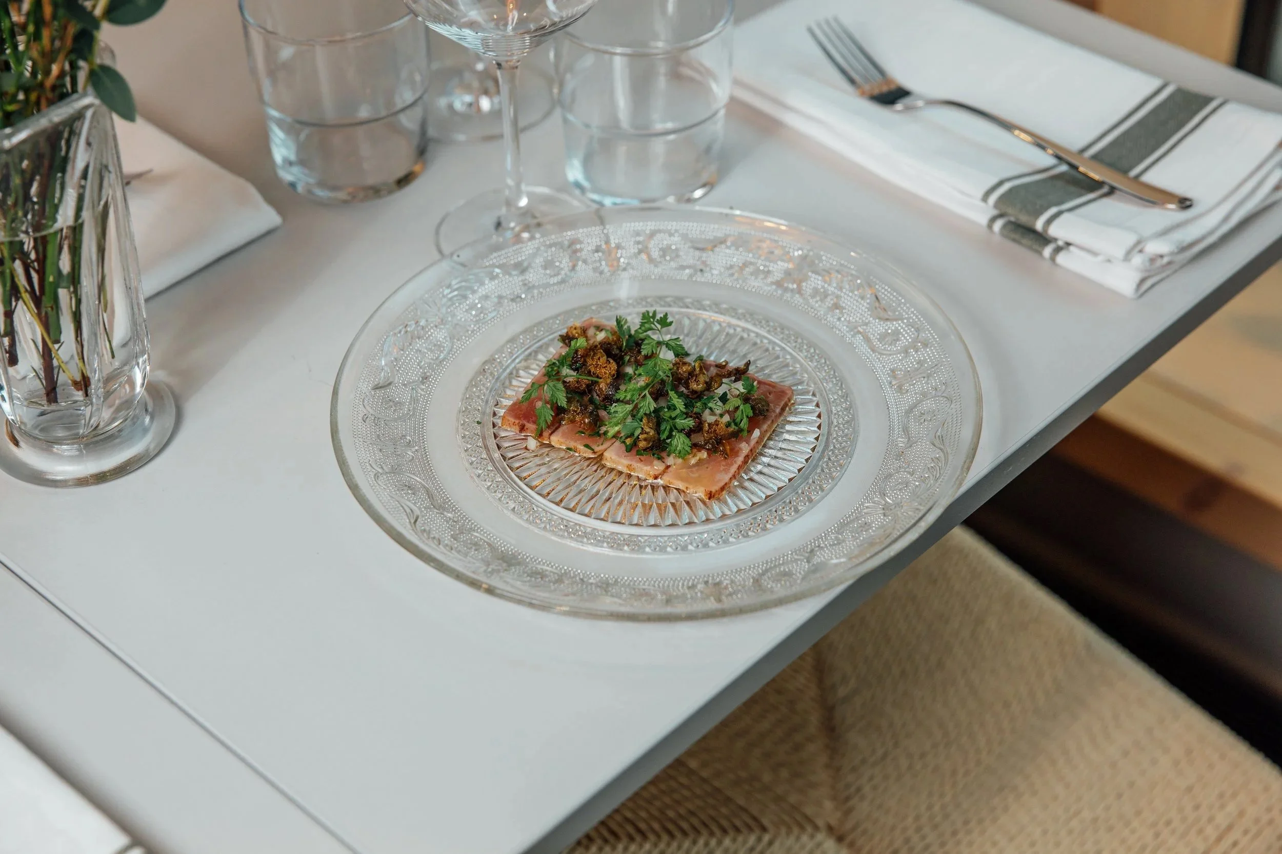 A glass plate with a serving of carpaccio garnished with herbs, on a white table setting with cutlery, glasses, and a floral arrangement.