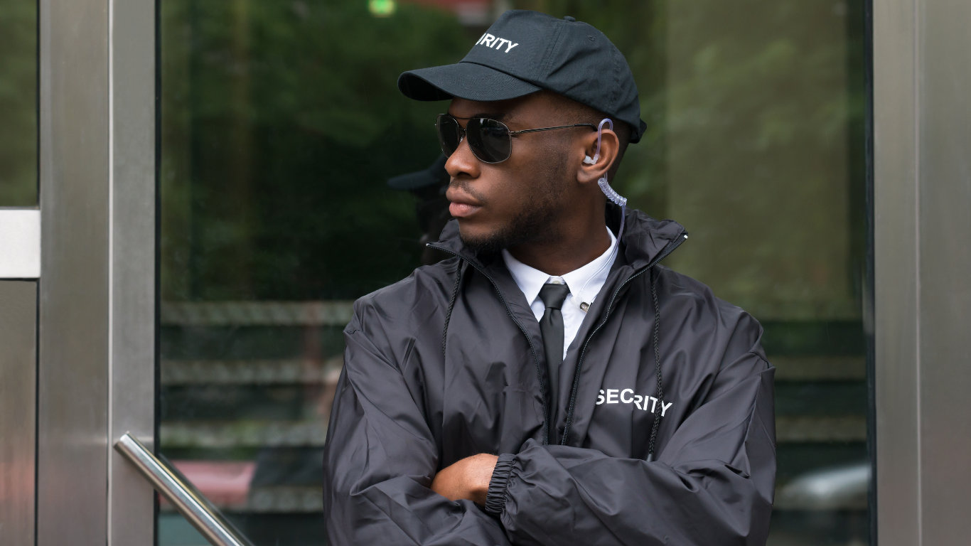 Security guard wearing sunglasses, black security uniform, and a cap, standing with arms crossed outside a building.