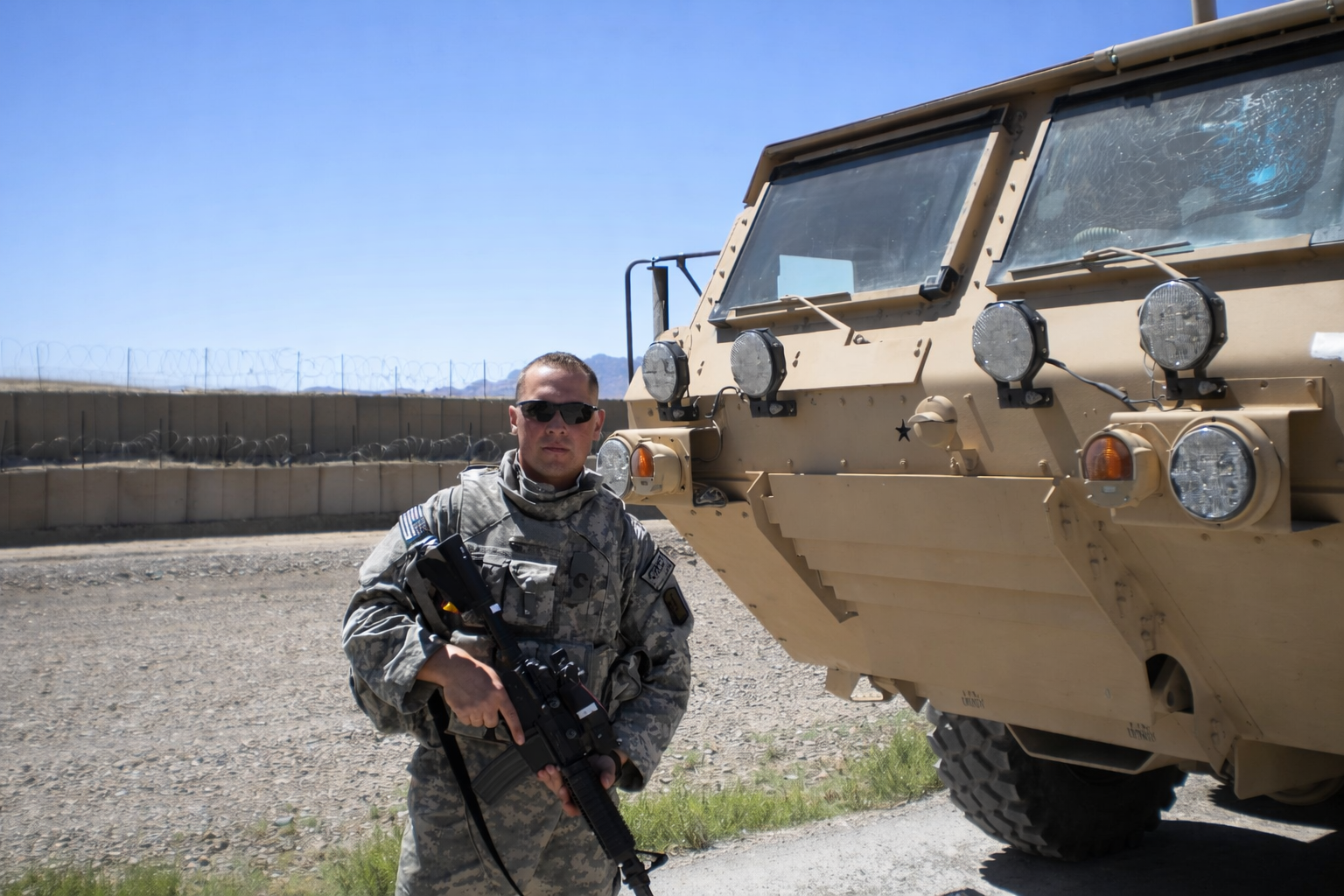 A soldier in camouflage uniform holding a rifle standing next to a military vehicle near a border fence under clear skies.